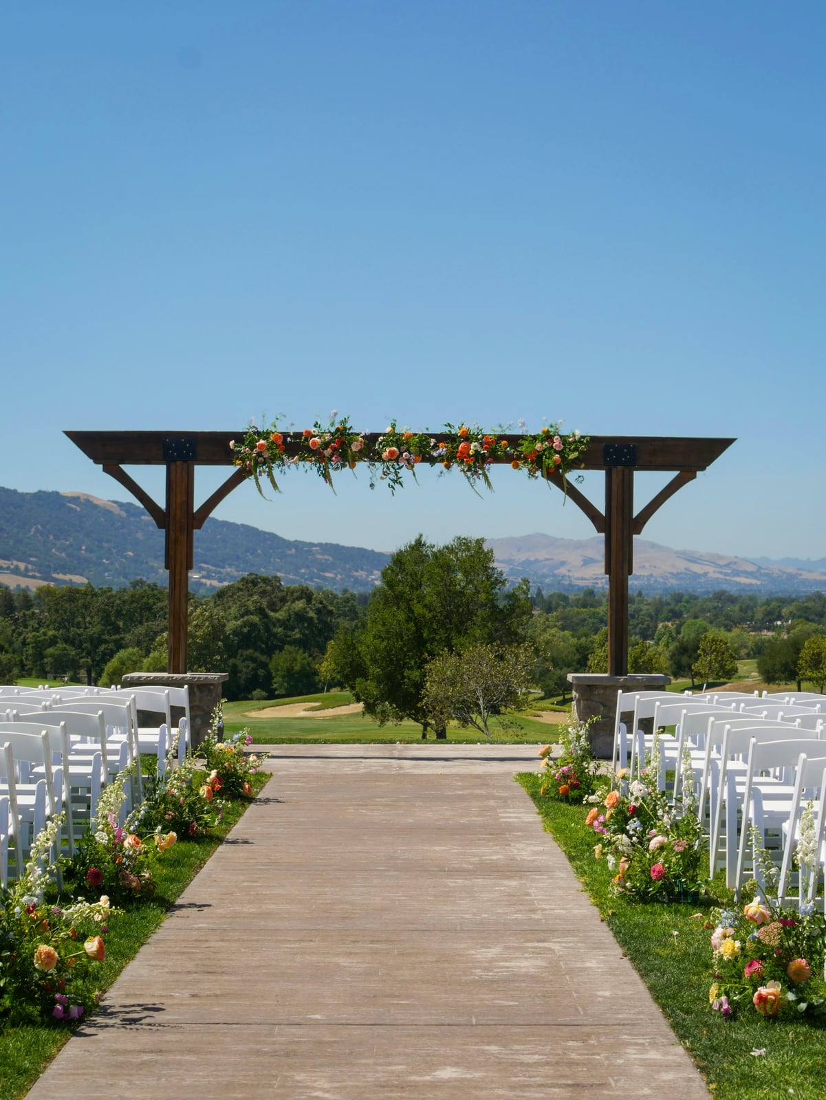 Outdoor ceremony aisle lined with white chairs beneath a wooden arch